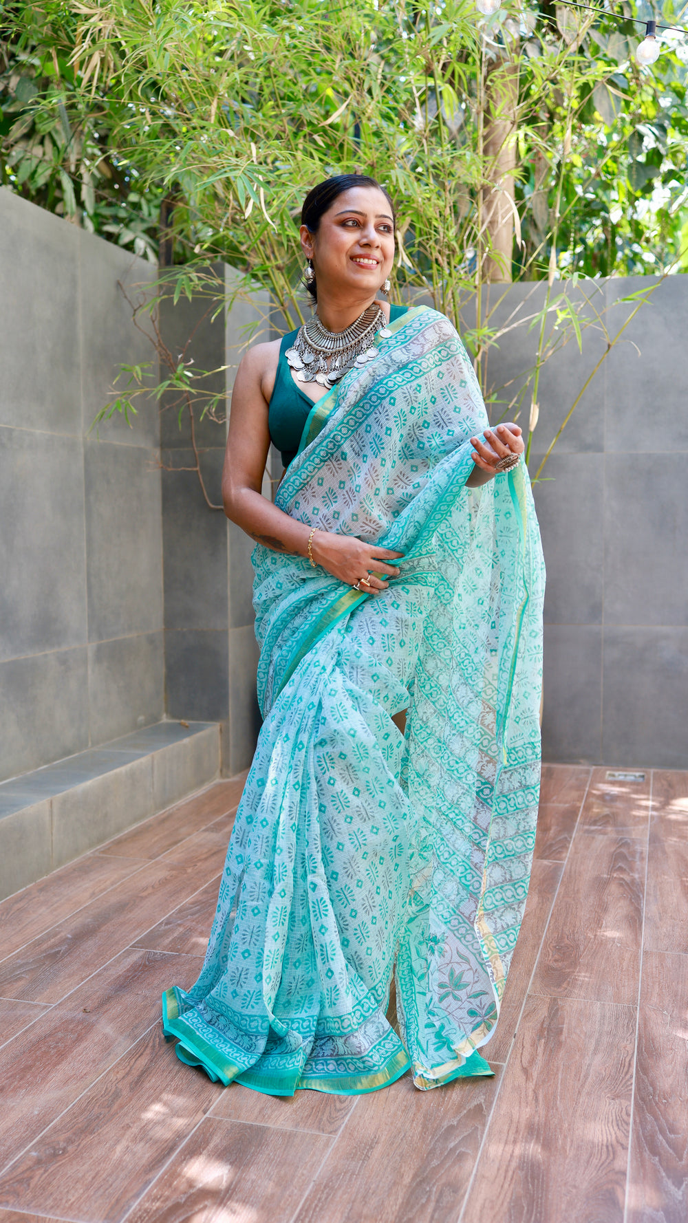Mint green and turquoise hand-block printed cotton saree with a sheer texture and teal border, worn by a smiling model styled with a sleeveless blouse and bohemian silver statement necklace.