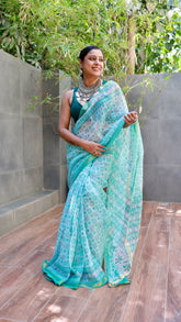 Mint green and turquoise hand-block printed cotton saree with a sheer texture and teal border, worn by a smiling model styled with a sleeveless blouse and bohemian silver statement necklace.