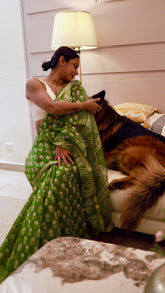 A woman draped in a green block-printed cotton-silk saree with a gold zari border, sitting on a couch and petting a fluffy German Shepherd dog in a cozy home setting.