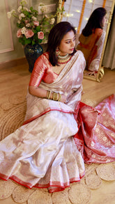 Full shot of a model sitting on a jute mat wearing an off-white Banarasi saree with a rich red zari woven border.