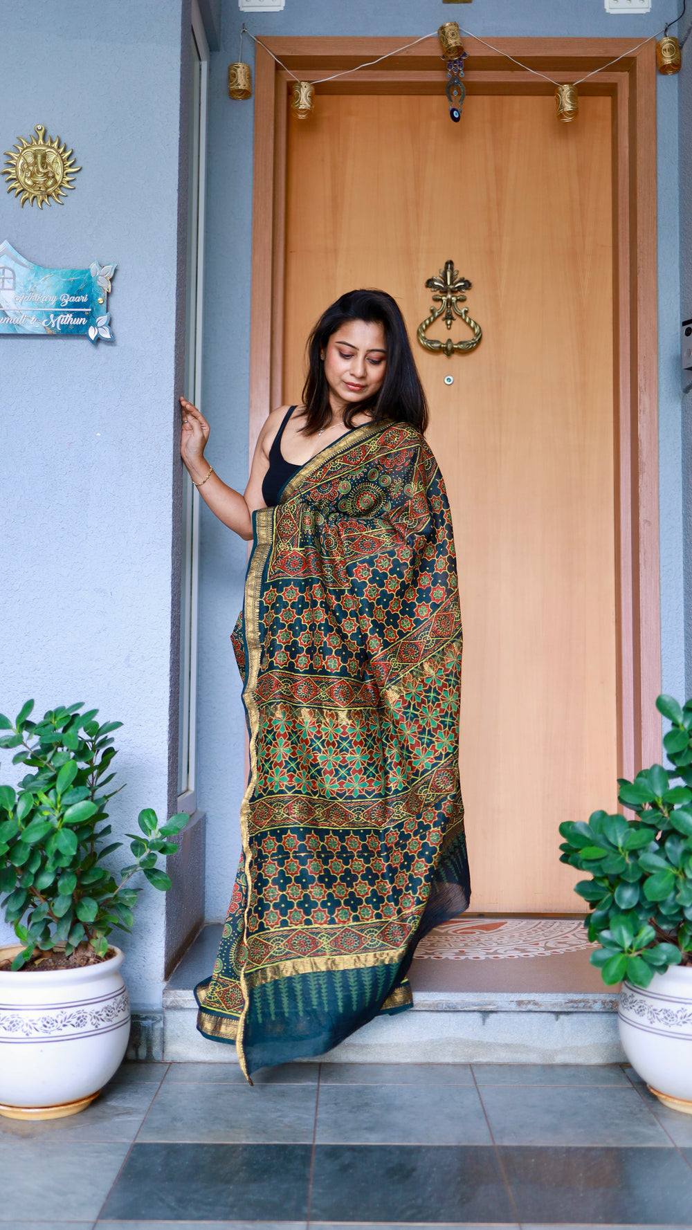 Woman draped in a teal Maheshwari Silk Saree with red and green Ajrakh block prints, posing near a wooden door.