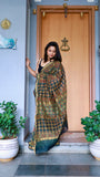 Woman draped in a teal Maheshwari Silk Saree with red and green Ajrakh block prints, posing near a wooden door.