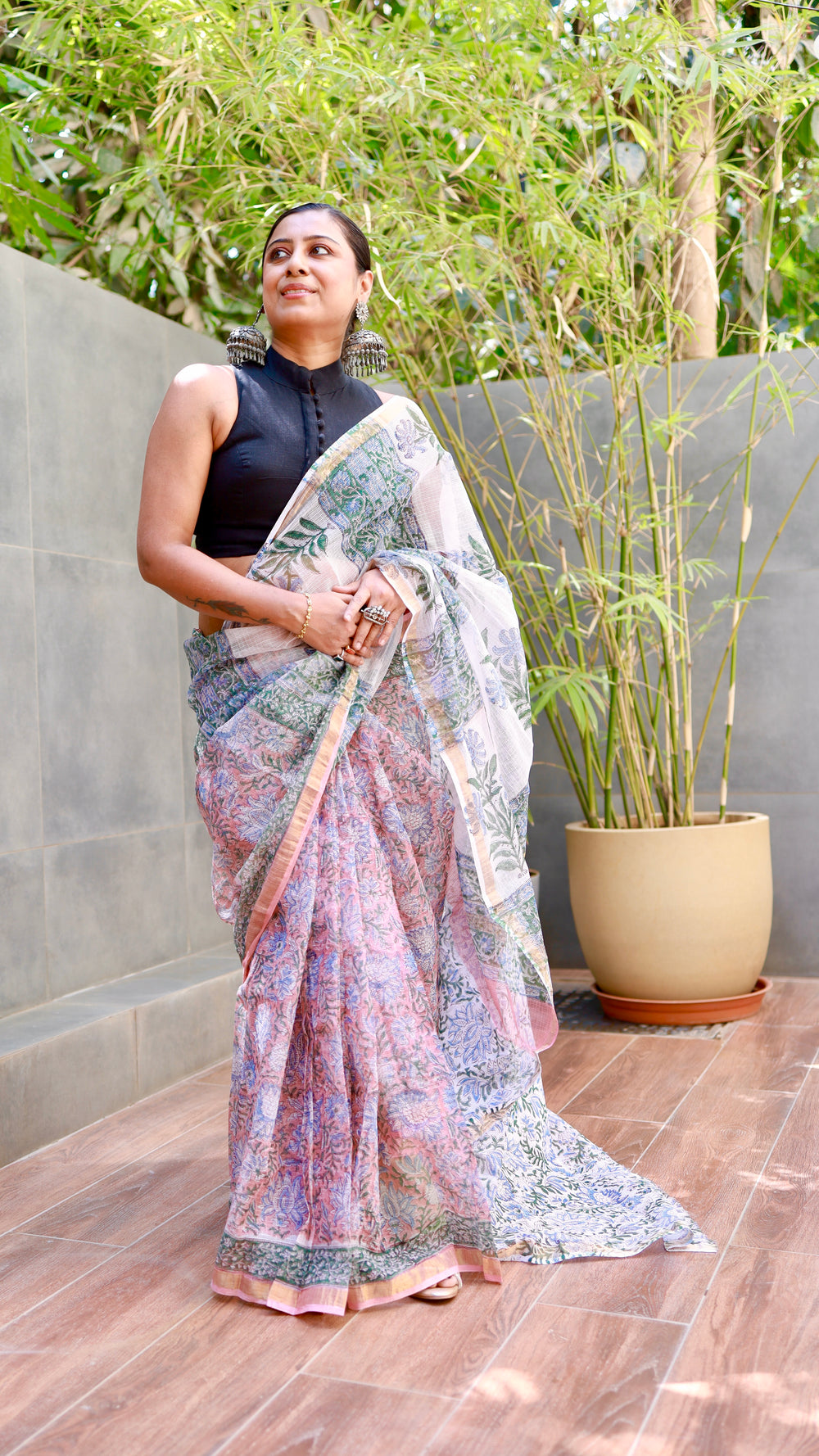 Woman wearing a Elegant hand-block printed pastel pink Kota Doria Cotton saree with a gold border, paired with a black sleeveless high-neck blouse and silver jhumka earrings.