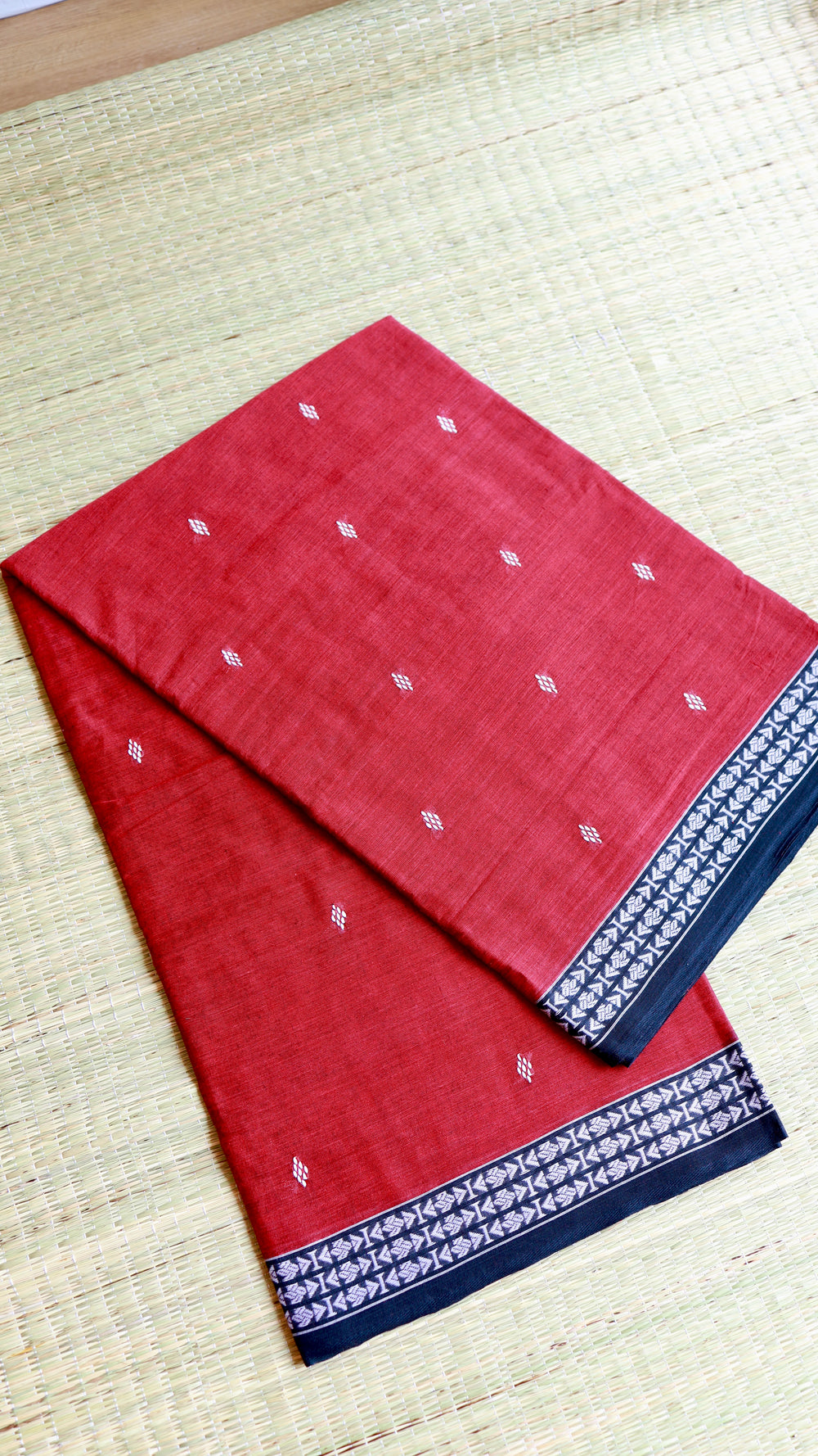 Neatly folded Red Dhonekhali Cotton Saree on a traditional mat showcasing the fabric texture and contrast border.