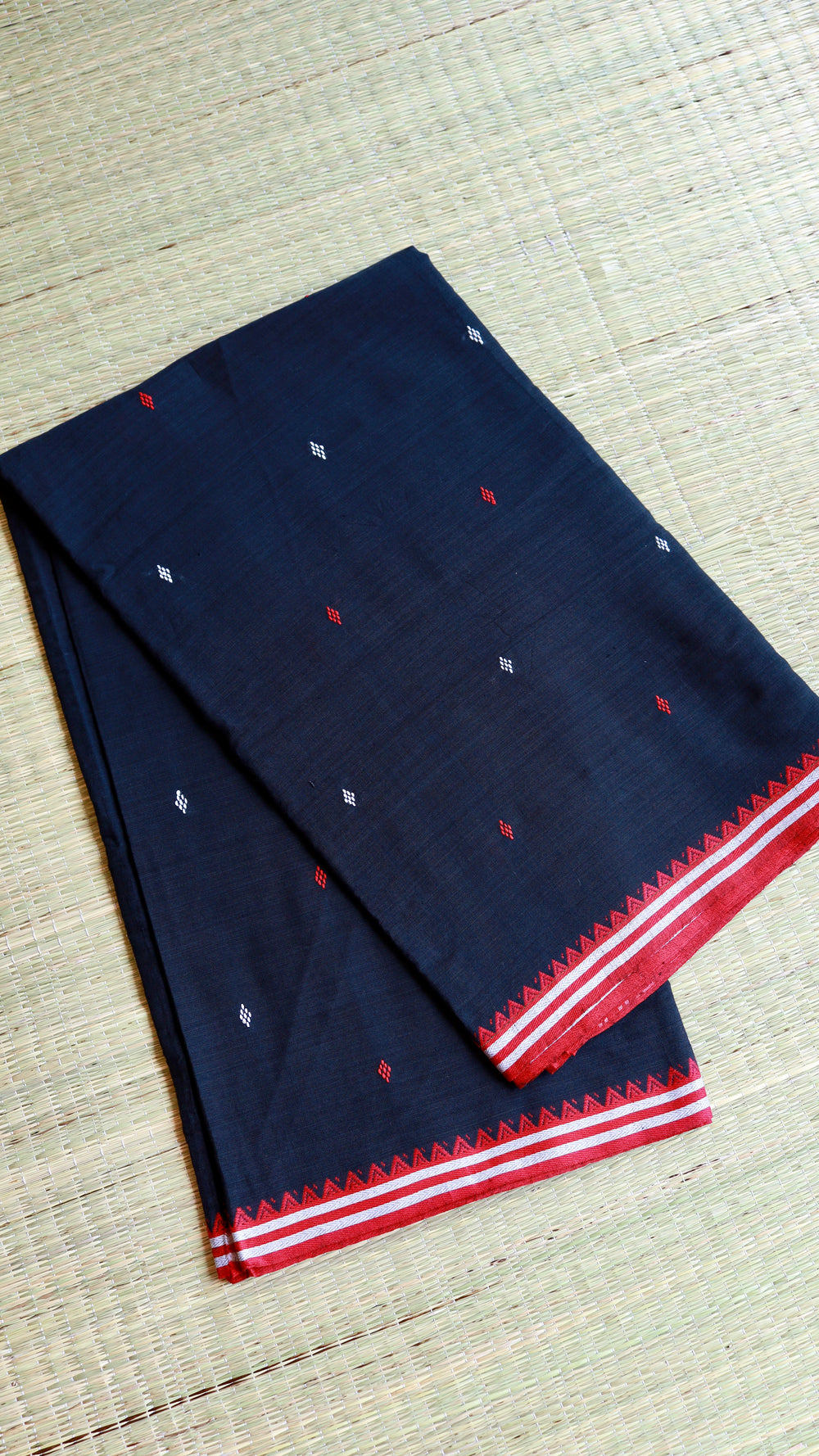 Flat lay of a Black Dhonekhali Handloom Cotton Saree on a traditional grass mat, showing red and white thread butti work