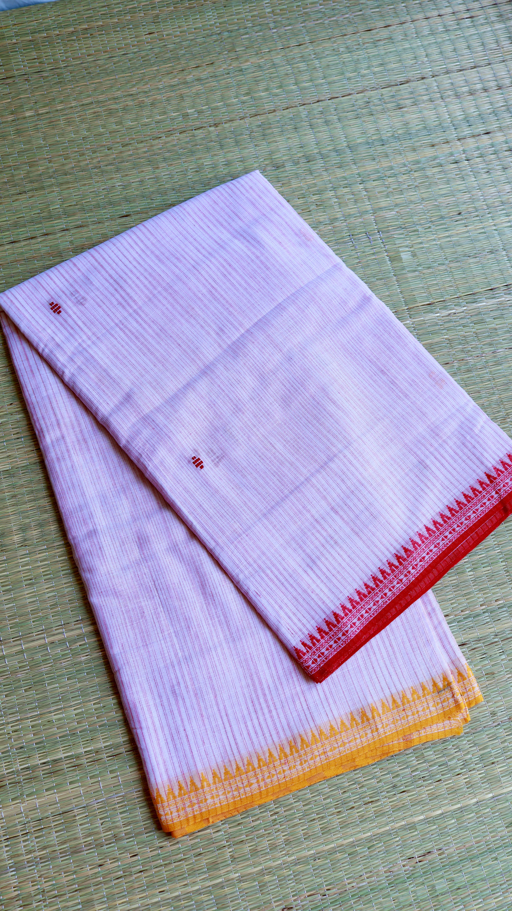 Folded Dhonekhali cotton saree on a grass mat highlighting the dual-toned red and mustard borders.