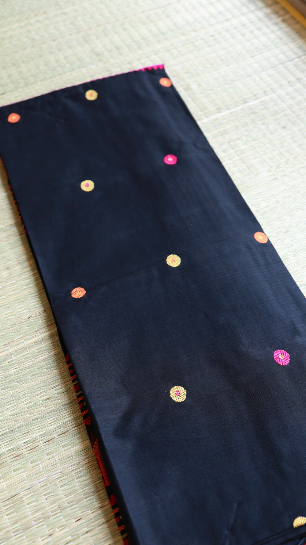 A neatly folded black Baluchari cotton saree showcasing all-over handwoven circular bootis in magenta pink, orange, and gold threads work
