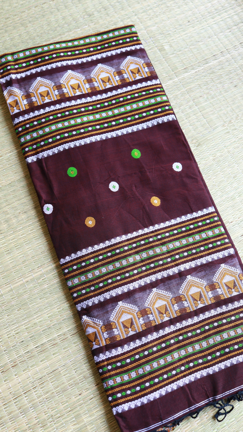 Flat-lay view of a folded coffee brown Baluchari cotton saree showing the stacked horizontal temple motifs on the pallu and the multicolored geometric borders.