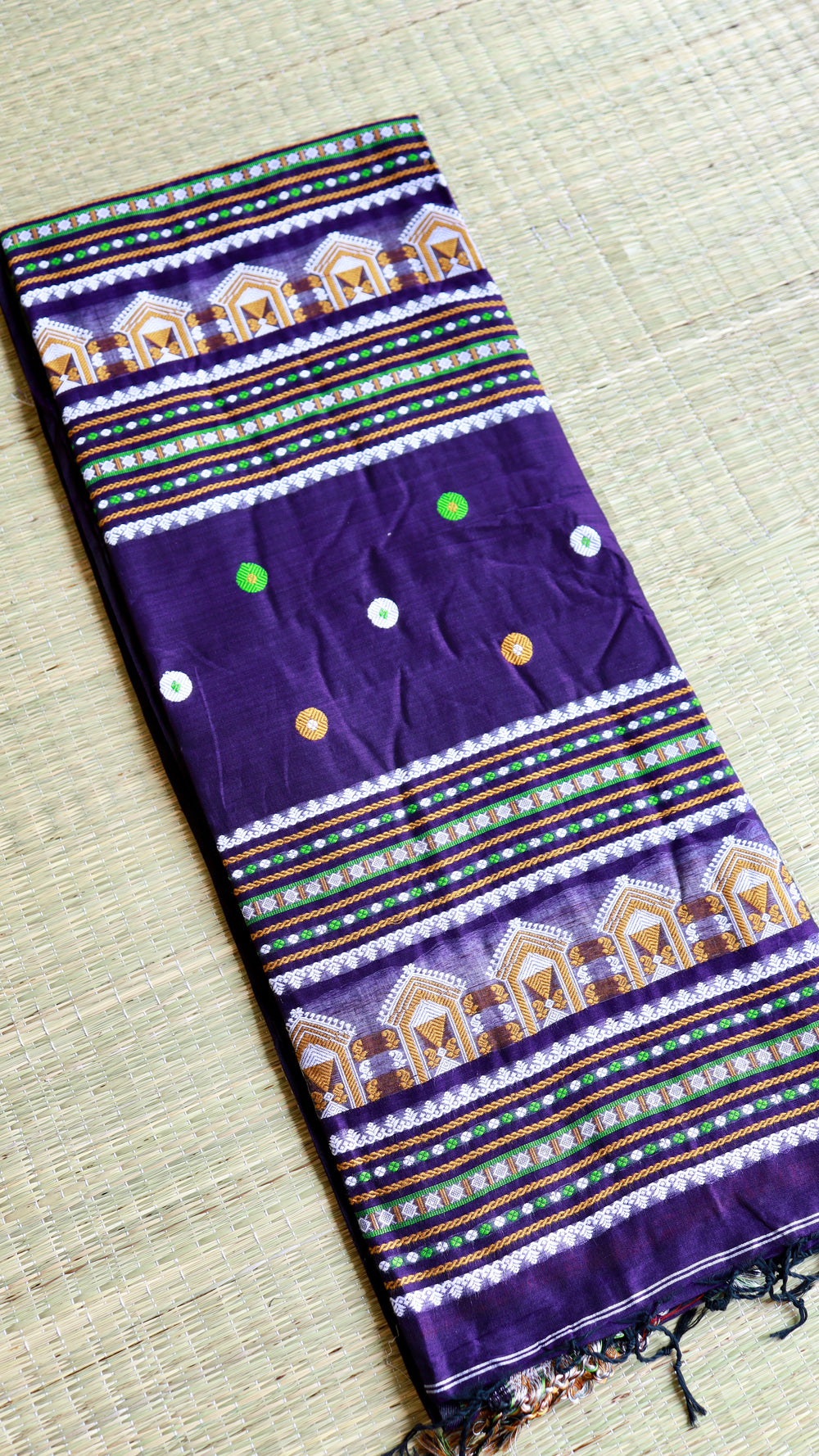 Top-down flat lay view of the folded deep purple Baluchari cotton saree on a woven mat, highlighting the symmetrical layout of the traditional hut patterns and vibrant borders.