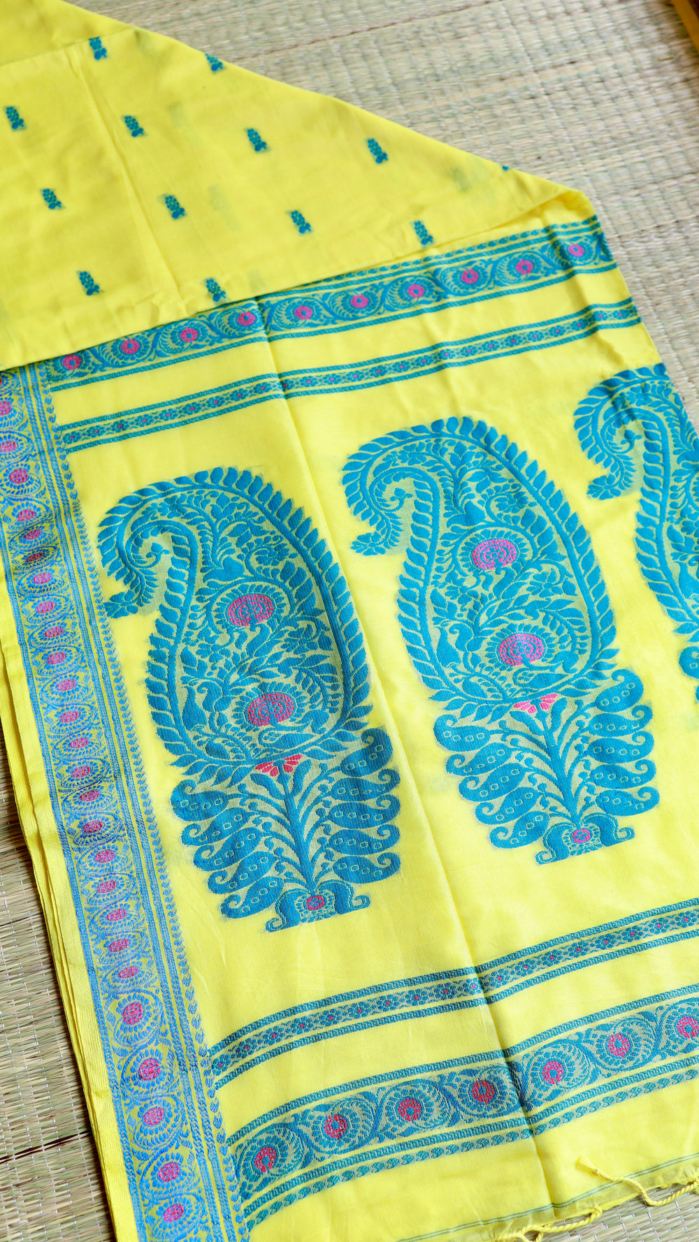 Overhead flat-lay view of the folded lemon yellow Baluchari cotton saree on a woven mat, revealing the grand paisley pallu design and matching blouse piece fabric layout.