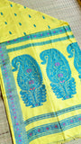 Overhead flat-lay view of the folded lemon yellow Baluchari cotton saree on a woven mat, revealing the grand paisley pallu design and matching blouse piece fabric layout.
