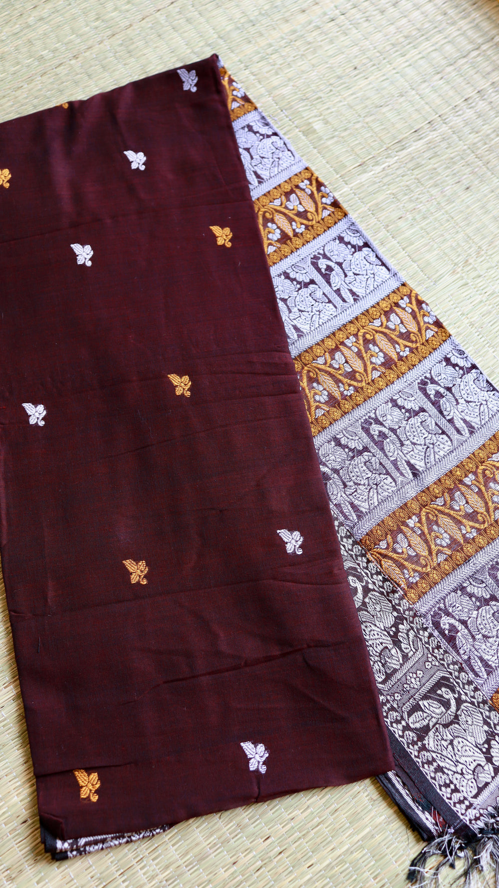A neatly folded coffee maroon Baluchari Cotton Saree resting on a natural grass mat, explicitly showing the stark contrast between the minimal body motifs and the heavily patterned narrative pallu.