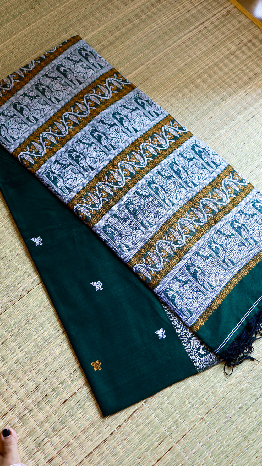 A neatly folded emerald green Baluchari Cotton Saree arranged flat on a traditional straw mat, clearly showing the contrast between the solid body weave and the ornate pallu