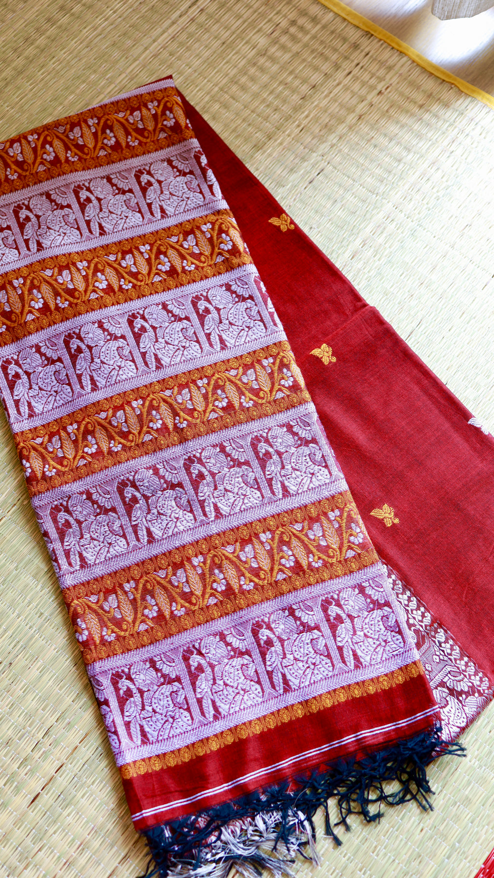 Close-up view of the folded pallu of a crimson red Baluchari Cotton Saree showing intricate white-silver figurative panels, mustard-orange floral borders, and hand-tied dark tassels.