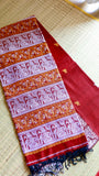 Close-up view of the folded pallu of a crimson red Baluchari Cotton Saree showing intricate white-silver figurative panels, mustard-orange floral borders, and hand-tied dark tassels.