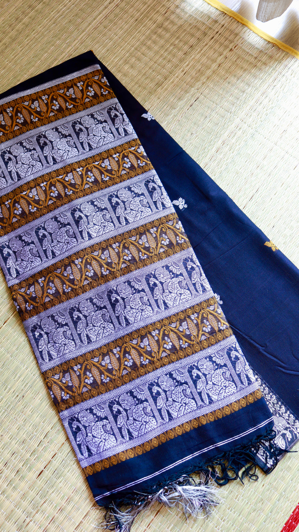 A folded close-up of the royal blue Baluchari Cotton Saree on a woven mat, highlighting the dense horizontal panels of silver and mustard zari mythological motifs on the pallu.