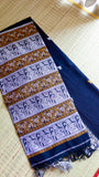 A folded close-up of the royal blue Baluchari Cotton Saree on a woven mat, highlighting the dense horizontal panels of silver and mustard zari mythological motifs on the pallu.