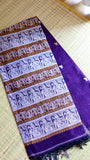 A neatly folded deep purple Baluchari cotton saree placed on a natural grass mat, explicitly detailing the elaborate woven horizontal panels of mythological patterns and fine fringe detailing at the edge.
