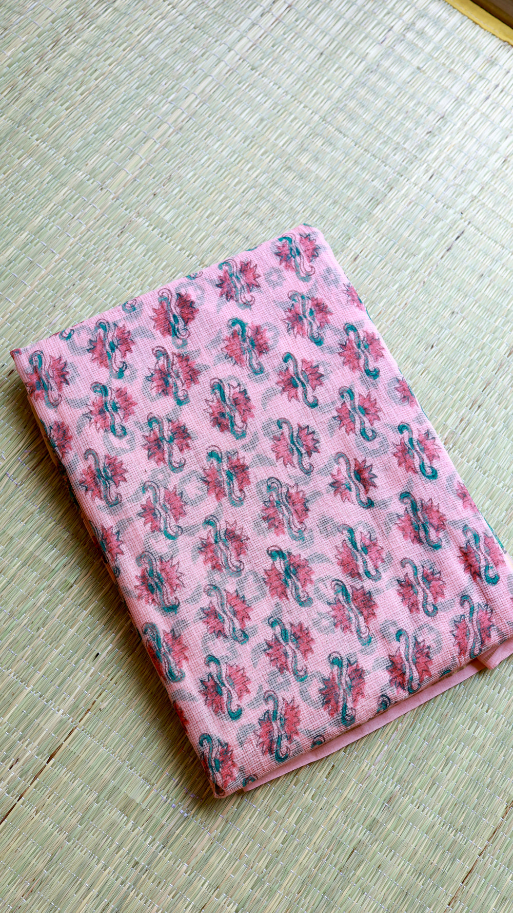 Neatly folded pink handblock Bagru printed Kota Doria pure cotton saree with its matching unstitched blouse piece resting on a woven natural grass mat.