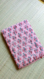 Neatly folded pink handblock Bagru printed Kota Doria pure cotton saree with its matching unstitched blouse piece resting on a woven natural grass mat.
