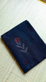 A neatly folded black Kota Doria pure cotton saree resting on a traditional grass mat, displaying a single hand-printed red floral block motif on the fabric surface.