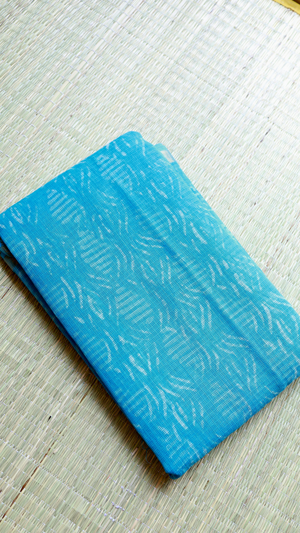 Flat lay image of the neatly folded sky blue Bagru printed Kota Doria pure cotton saree resting on a natural straw mat, showcasing the geometric handloom textures.
