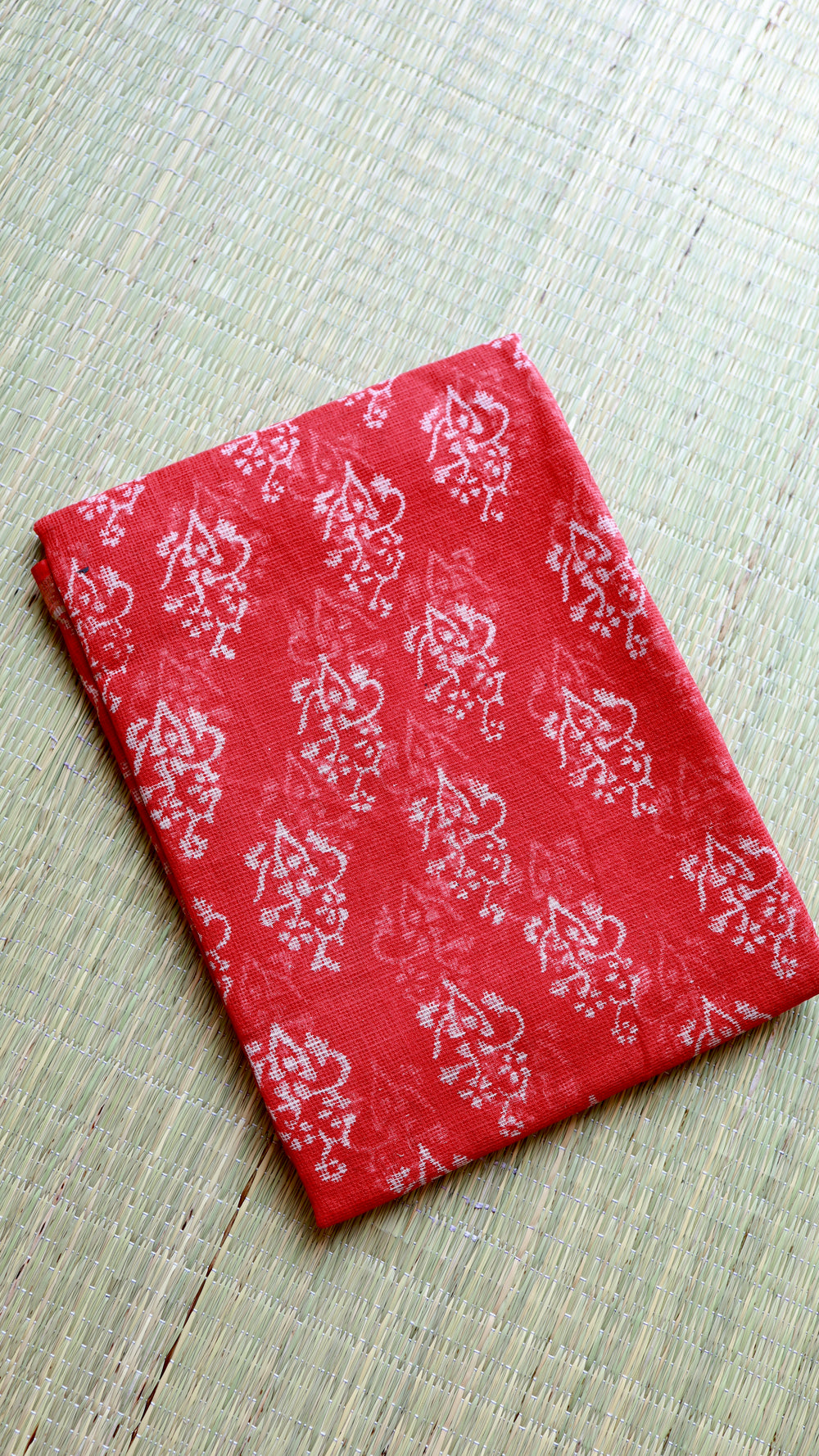 A neatly folded red Handblock Bagru printed Kota Doria cotton saree with matching blouse piece fabric, displayed flat on a natural woven grass mat.