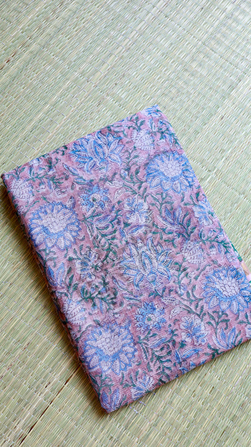 Neat rectangular fold of the pure cotton pink Kota Doria saree featuring intricate blue and white floral handblock prints on a natural grass mat.