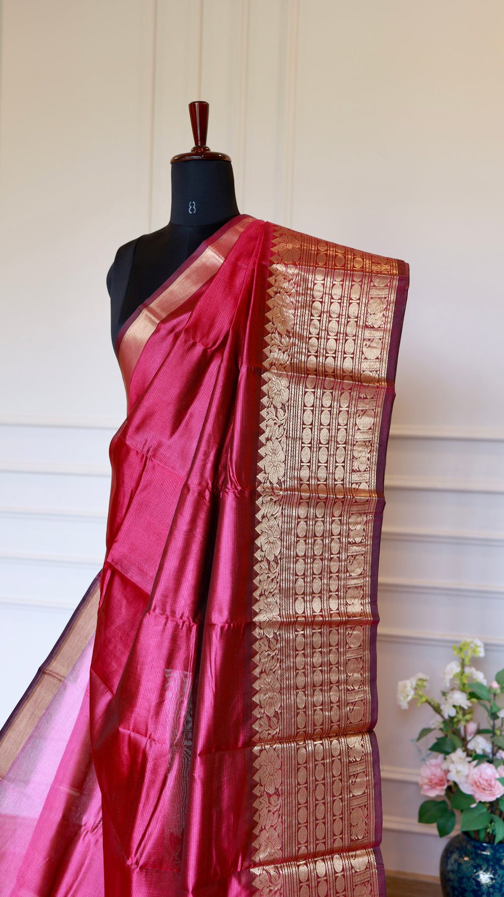 A deep Rani pink silk saree with an ornate gold zari border, draped on a black mannequin torso against a cream paneled wall.