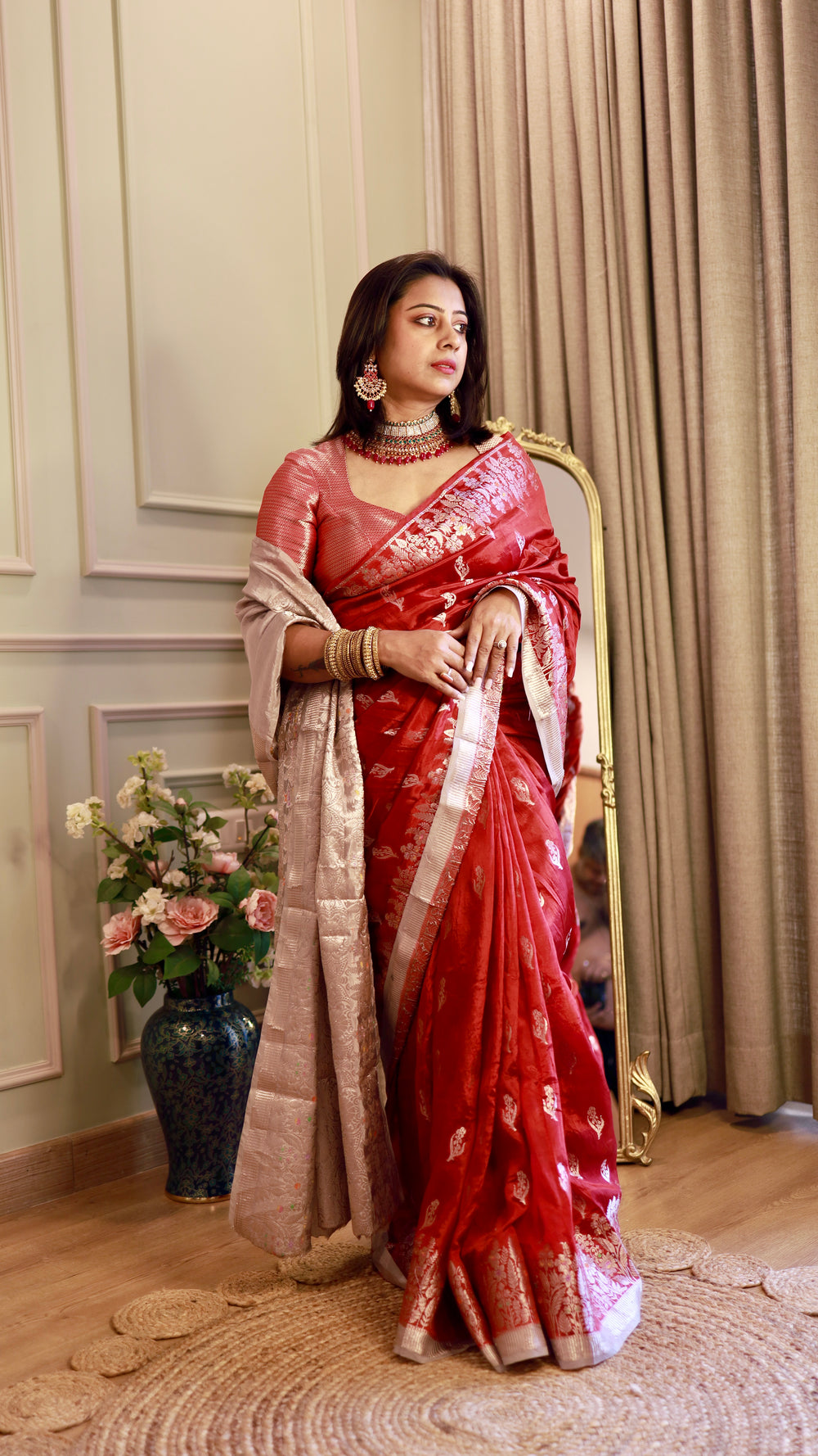 Model standing gracefully in a Crimson Red Georgette Banarasi Saree featuring silver zari motifs and an draped tissue pallu in a classic indoor setting.