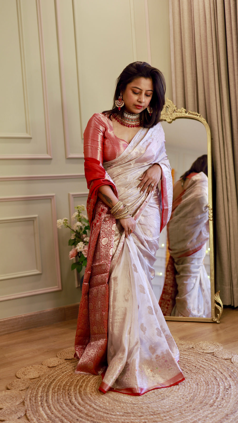 Full-length portrait of a model standing near a mirror showcasing the fluid drape of the off-white and crimson Banarasi saree.
