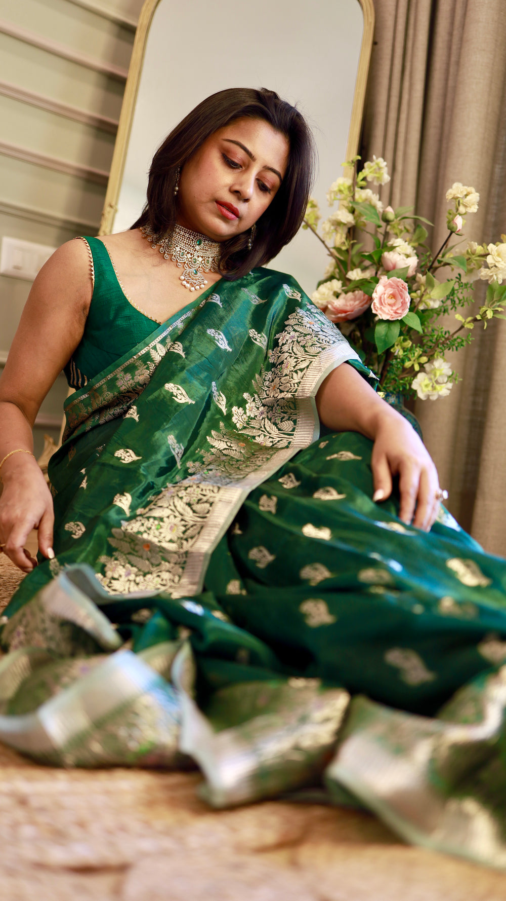 Woman wearing an emerald green georgette Banarasi saree with silver zari floral bootis sitting gracefully near a mirror.