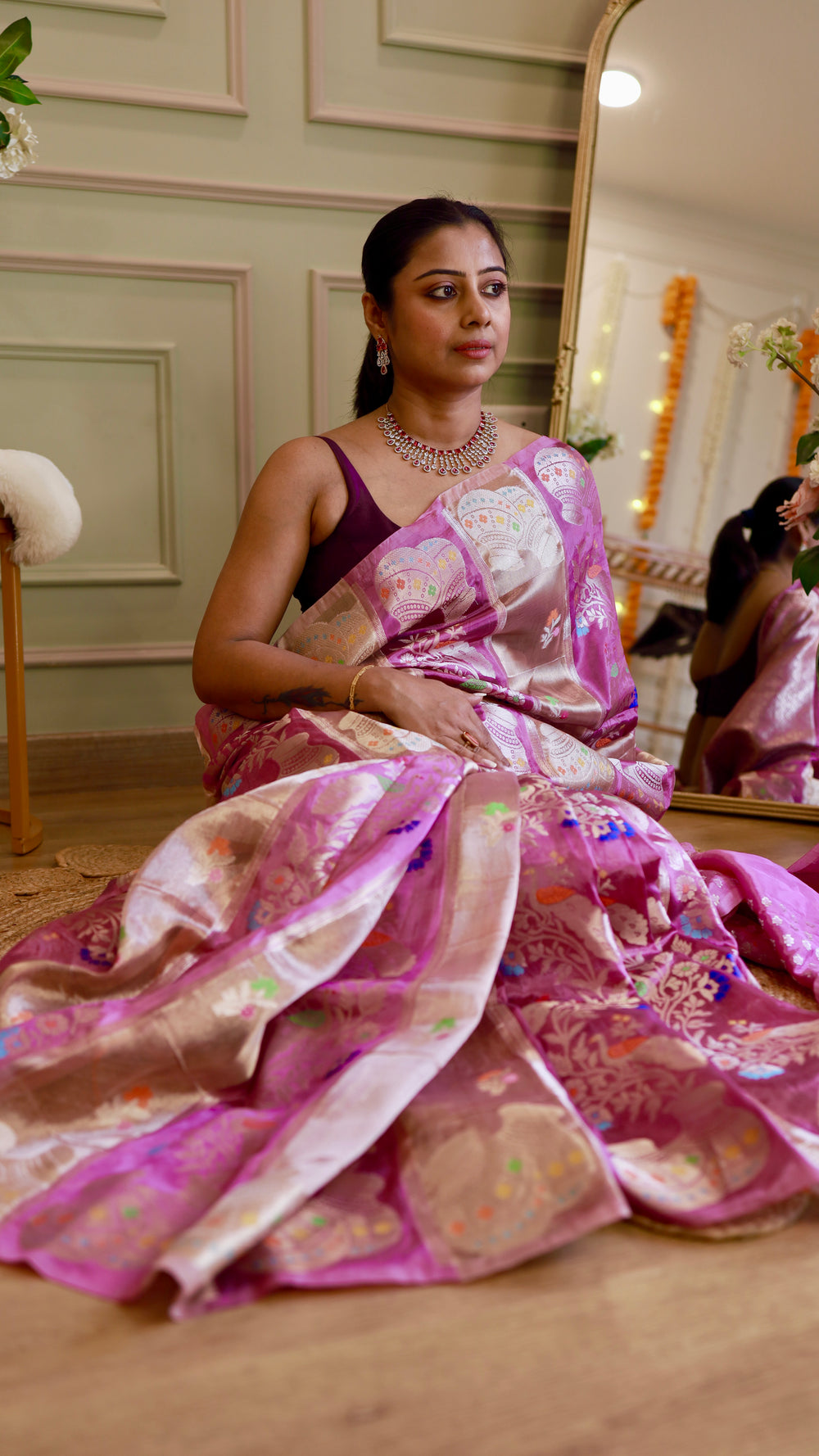Seated floor shot highlighting the silk sheen and colorful Meenakari peacock weave on the pallu.