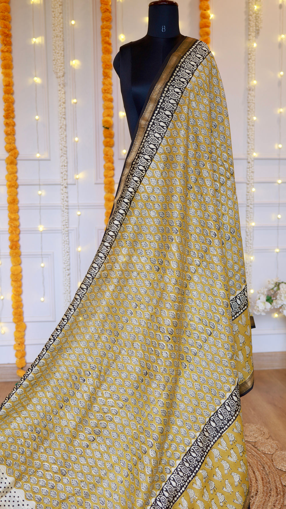 Mustard yellow hand block printed Maheshwari silk saree with black paisley woven zari border draped on a mannequin.