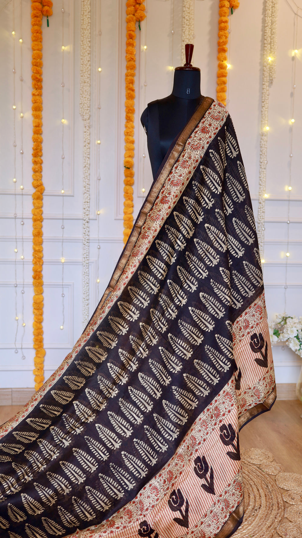 Full pleat and drape view of charcoal black Maheshwari silk saree with white leaf block prints, ornate red pallu border, and gold zari hem.