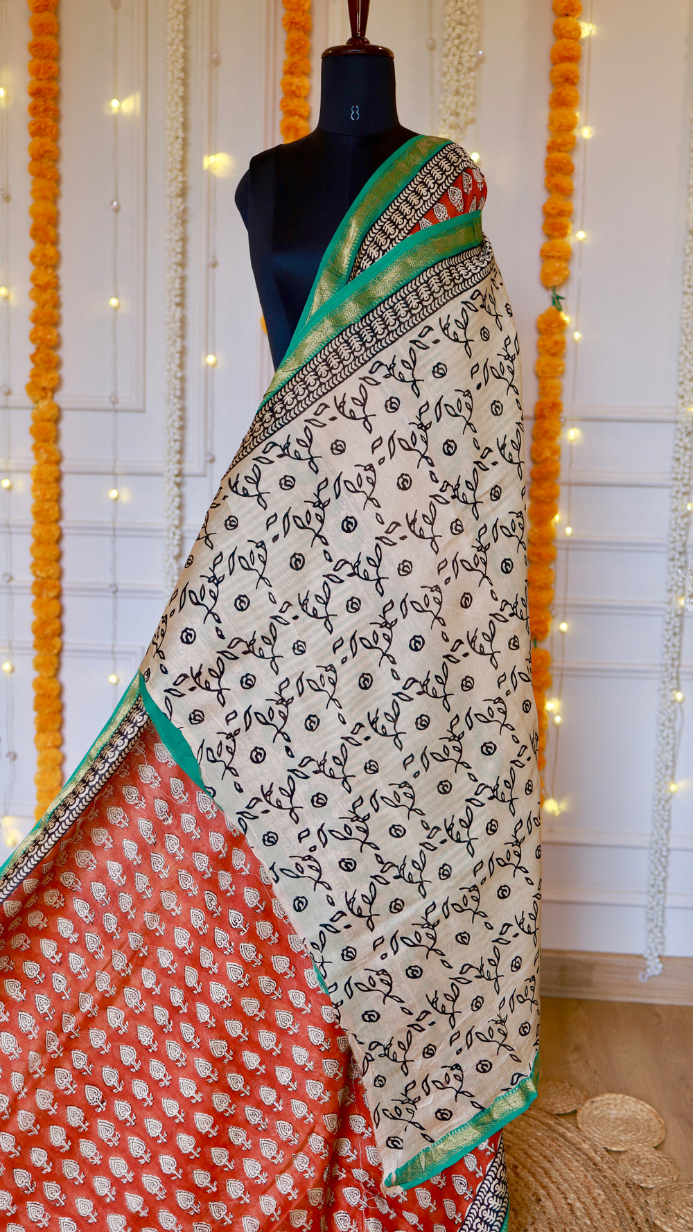 Rust Red Maheshwari Silk Saree draped on a mannequin showing off-white floral block-printed pleats and emerald zari border.
