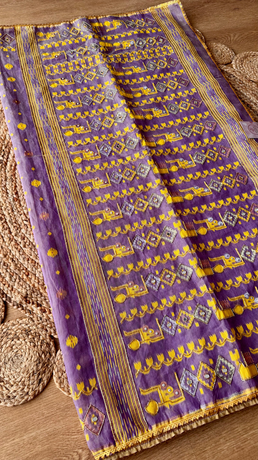 Close-up of handwoven lavender Kardana Jamdani saree pallu featuring mustard yellow motifs, glass beadwork, and mirror embellishments on a woven jute mat background.