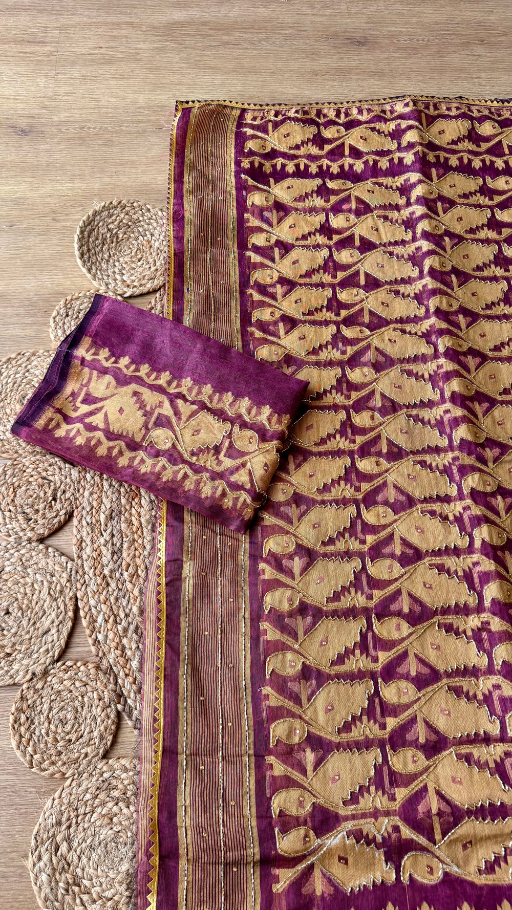 Top angle shot of wine purple Kardana Jamdani saree with matching unstitched blouse piece laid over textured jute mat.