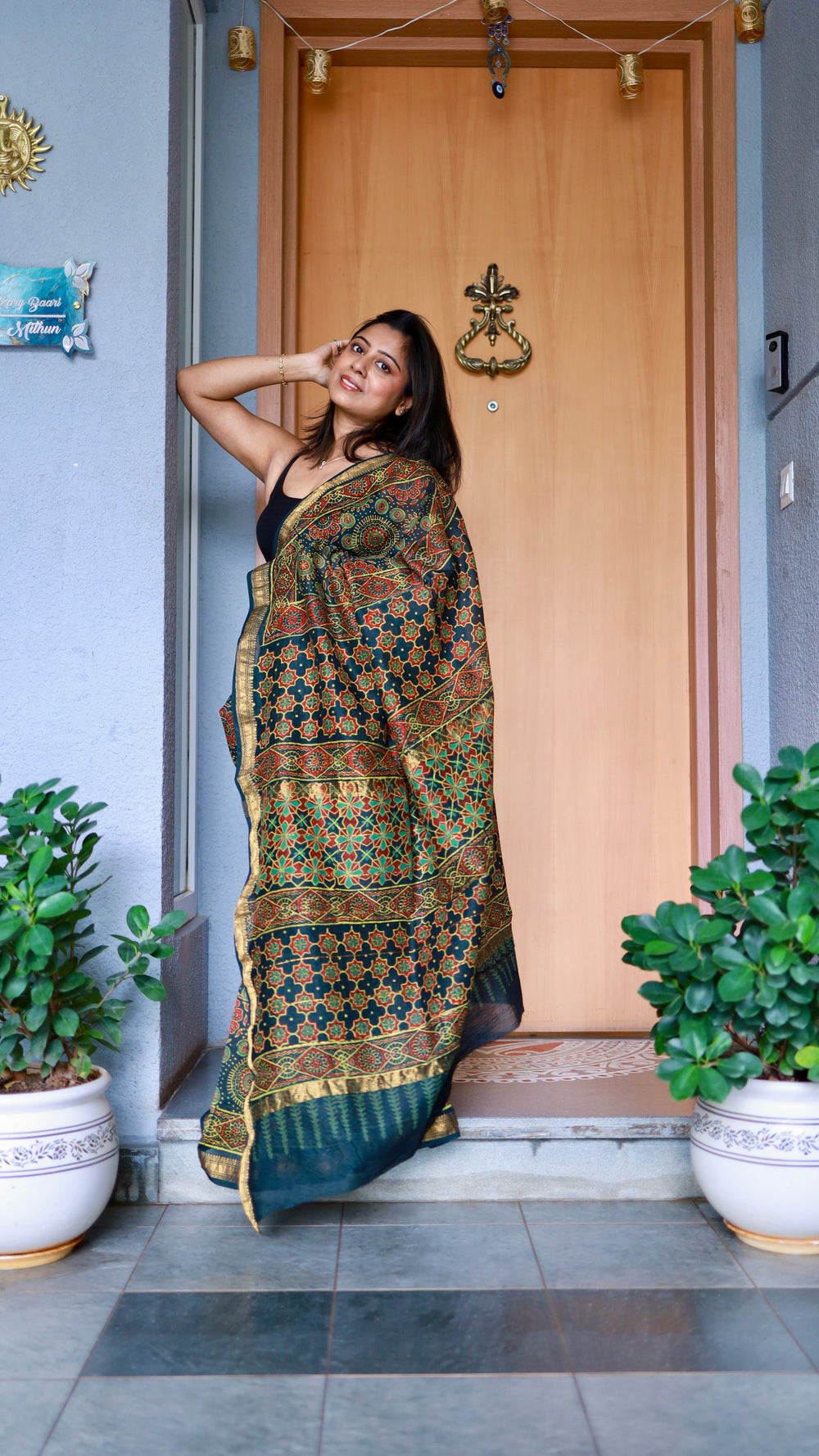 Woman draped in a teal Maheshwari Silk Saree with red and green Ajrakh block prints, posing near a wooden door.