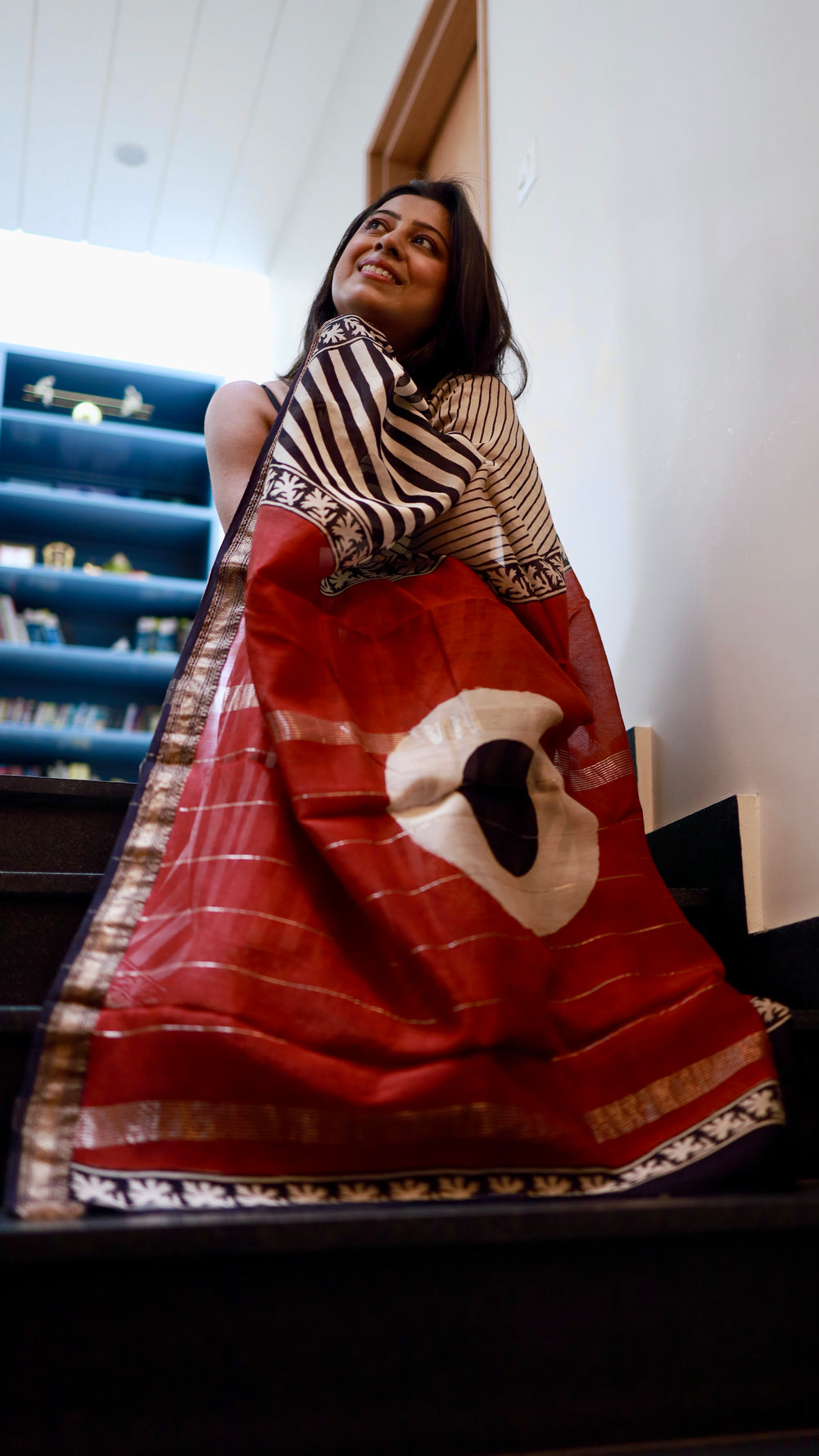 A woman smiling, draped in a Titafu Maheshwari Silk Saree, highlighting the flow of the fabric on a staircase.